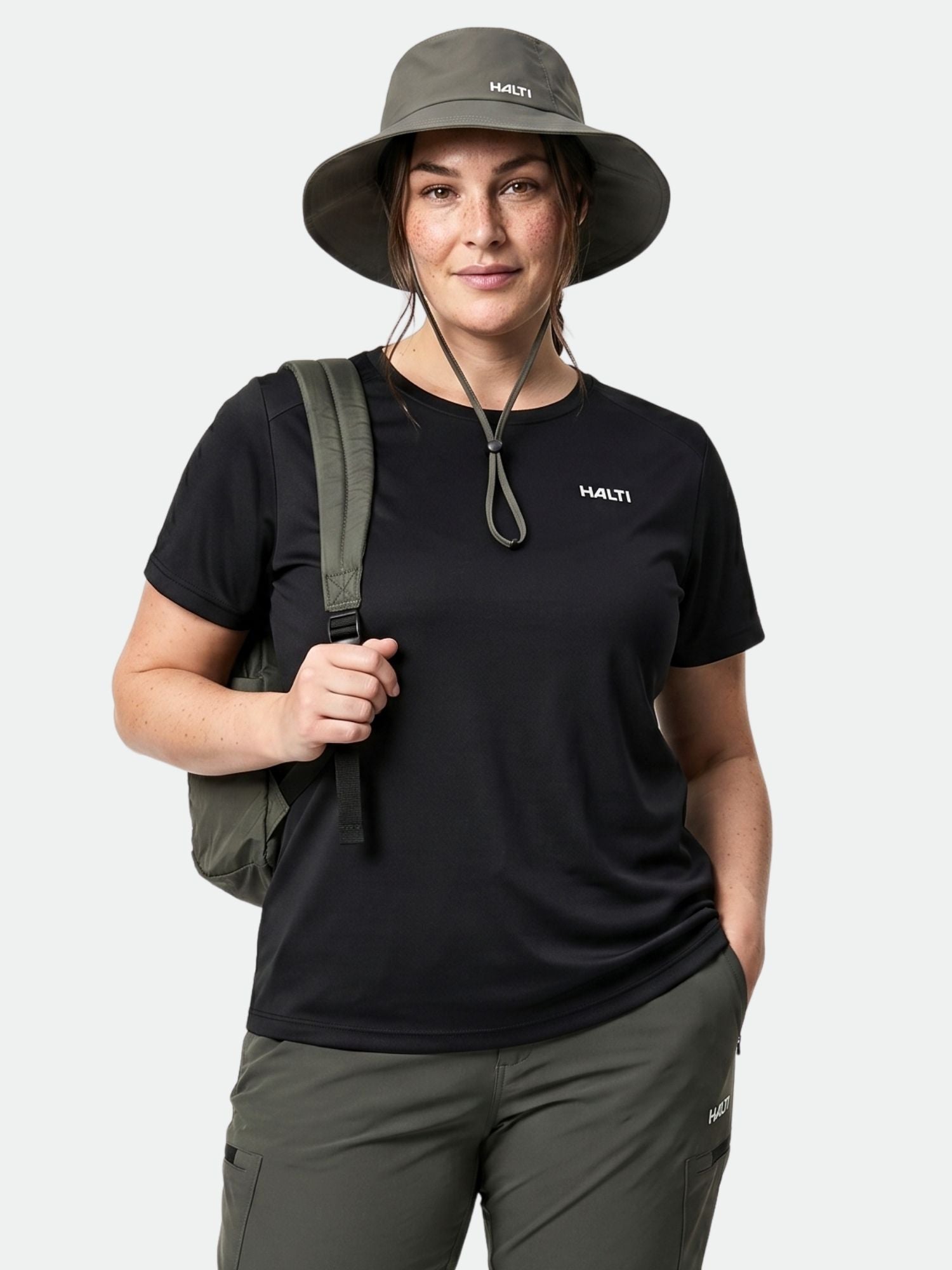 Eine Frau in Outdoor-Kleidung trägt vor einem schlichten Hintergrund ein „Halti Salves T-Shirt Damen Plus“, einen Hut mit breiter Krempe und einen Rucksack.