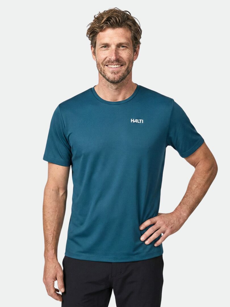 Mann lächelnd in einem tealfarbenen Halti Salves T-Shirt Herren, gepaart mit einer schwarzen Hose vor einem schlichten Hintergrund.