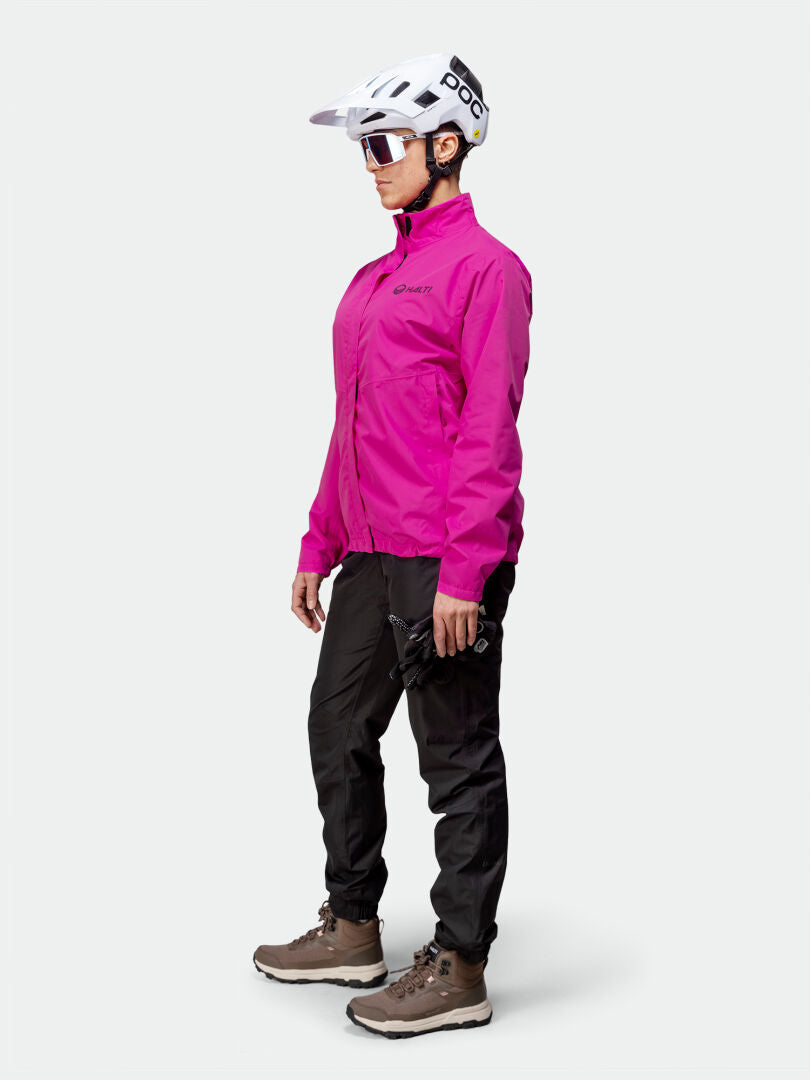 Eine Frau trägt die Halti Triant DX Fahrradjacke in Pink mit reflektierenden Details, schwarze Hosen und Wanderschuhe.
