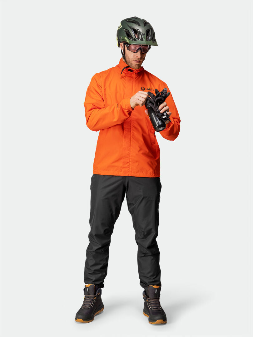 Eine Person in einer Halti Triant DX Radjacke für Herren, dunkler Hose und Helm steht da und rückt schwarze Handschuhe zurecht.