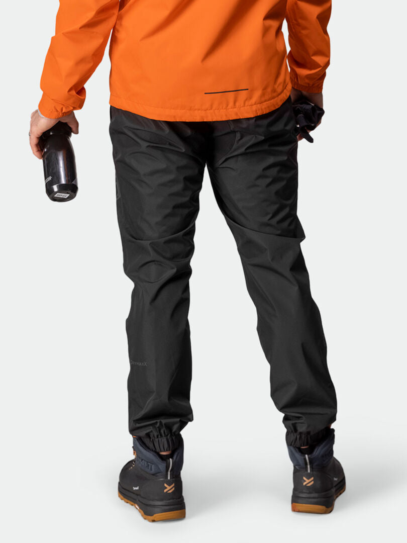 Person in orangefarbener Jacke, Halti Triant DX Fahrradhose für Herren, Wanderstiefeln, hält eine Wasserflasche mit dem Rücken zur Kamera.