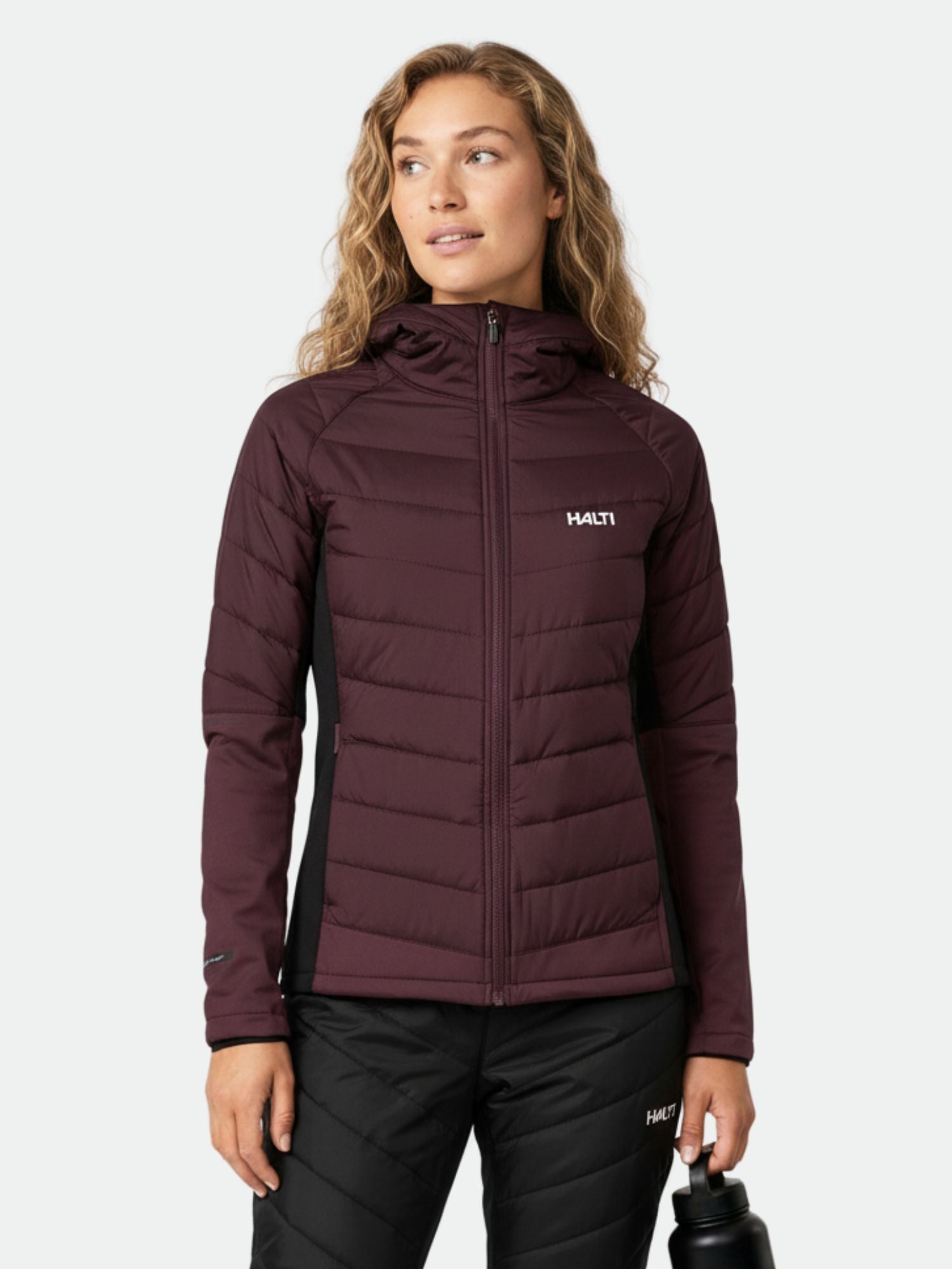 Eine Person in einer kastanienbraunen Halti Hanki 2 Hybrid Langlaufjacke Damen hält eine schwarze Wasserflasche vor einem schlichten Hintergrund.