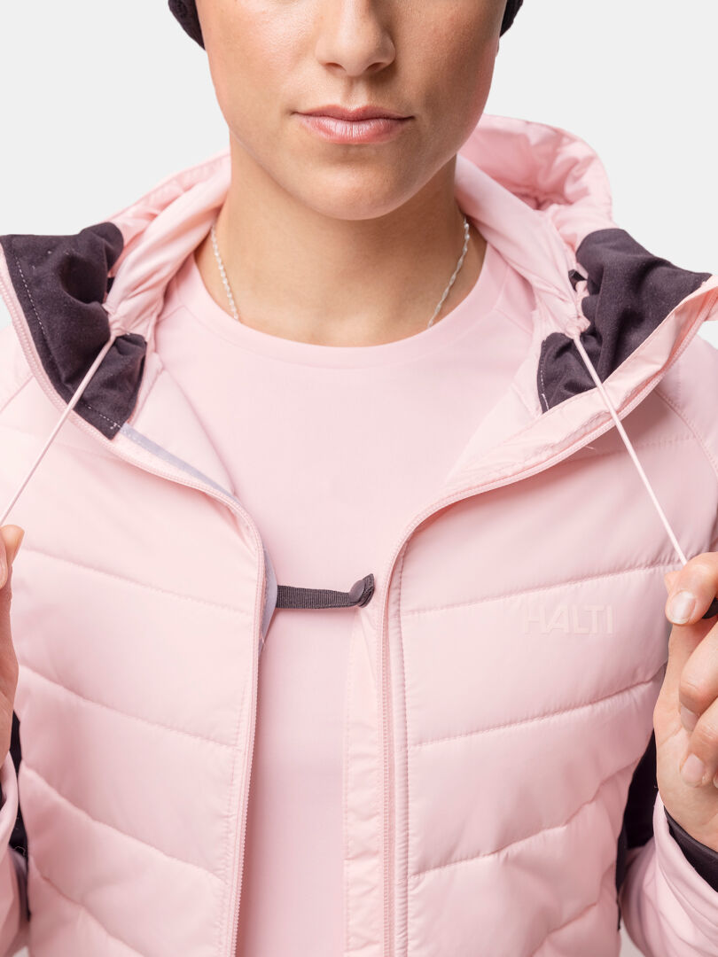 Frau in Halti Hanki 2 Hybrid Langlaufjacke hält Skistöcke, trägt pinke Jacke, schwarze Hose, Handschuhe, Mütze.