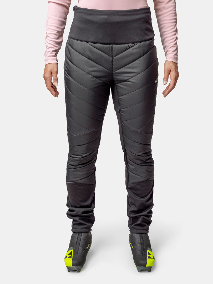 Eine Person trägt eine schwarze Halti Hanki 2 Hybrid Langlaufhose Damen, ein rosafarbenes langärmeliges Oberteil und schwarz-gelbe Skistiefel, mit Blick nach vorne.