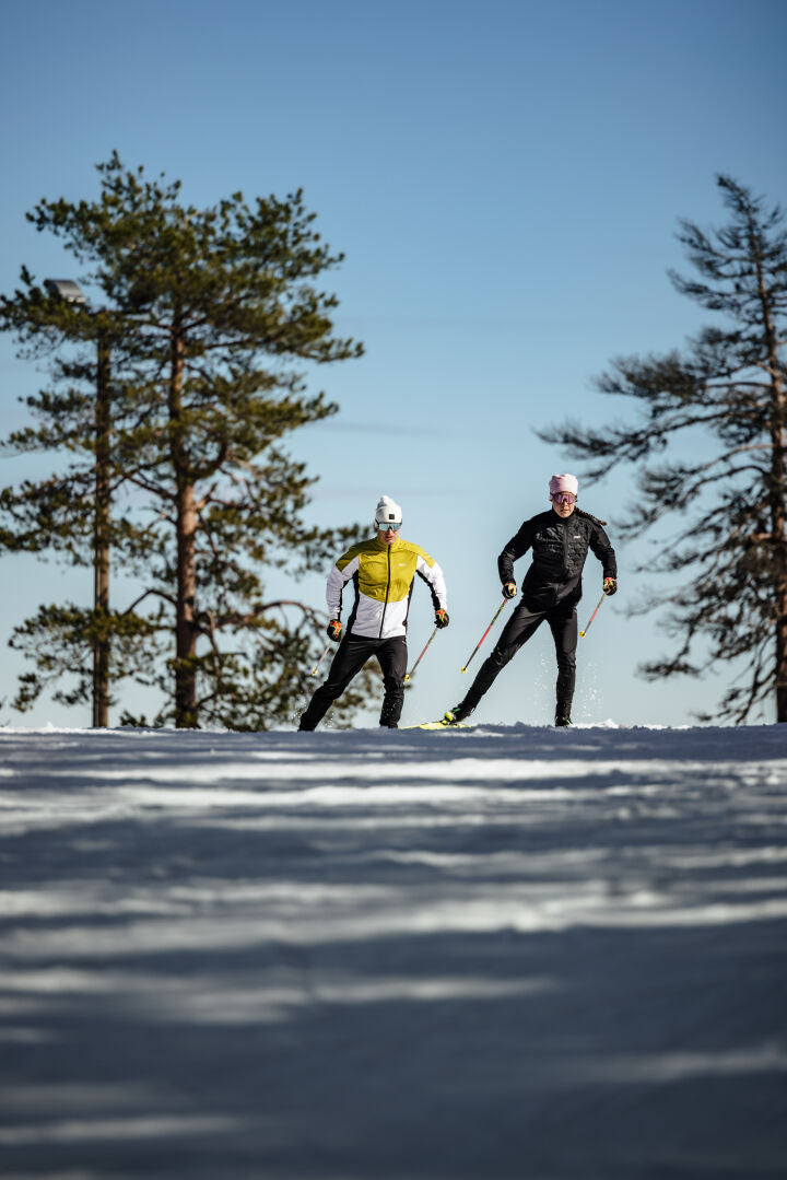 Zwei Männer in Halti Framover Langlaufjacke Herren beim Langlauf auf einer verschneiten Loipe unter strahlend blauem Himmel.