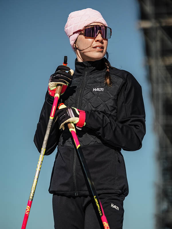 Mit der Halti Framover Hybrid Langlaufjacke Damen trägt eine Frau einen rosa Hut und eine Sonnenbrille im Freien mit Skistöcken.