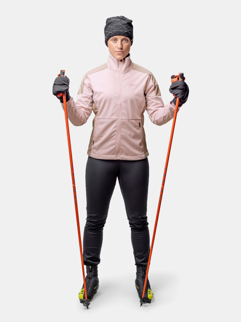 Eine Person in einem Halti Huuru Langlaufanzug Damen steht mit orangefarbenen Skistöcken und Skiern vor einem schlichten weißen Hintergrund.