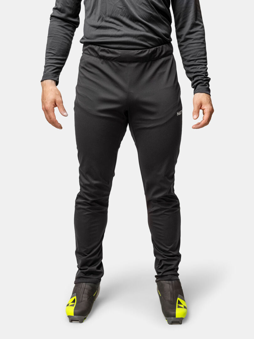 Eine Person in Halti Framover Langlaufhose Herren, einem langärmeligen schwarzen Hemd und schwarzen Schuhen mit gelben Akzenten steht aufrecht.