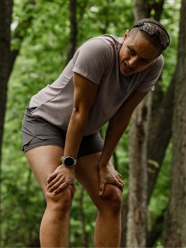 Frau in Halti Oulanka T-Shirt Damen beugt sich mit den Händen auf den Knien in einem Wald und holt Luft.