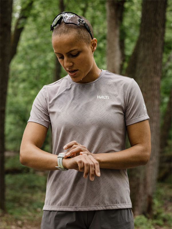 Frau in Halti Oulanka T-Shirt Damen prüft Smartwatch im Wald, Sonnenbrille auf dem Kopf liegend.