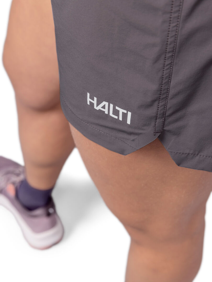 Nahaufnahme einer Person in Halti Uurto Shorts Damen (grau) und lilafarbenen Sportschuhen, die auf einem weißen Hintergrund steht.