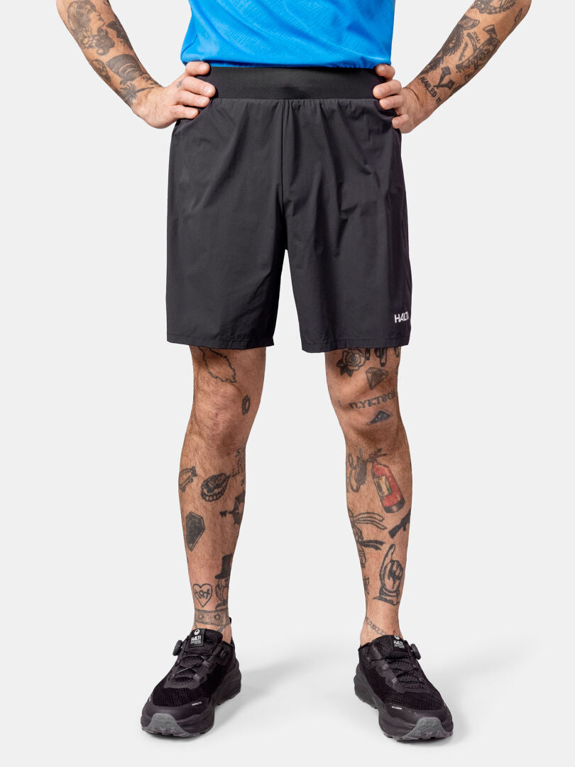 Eine Person in Halti Uurto Shorts Herren und schwarzen Schuhen steht mit den Händen in den Hüften und zeigt tätowierte Beine.