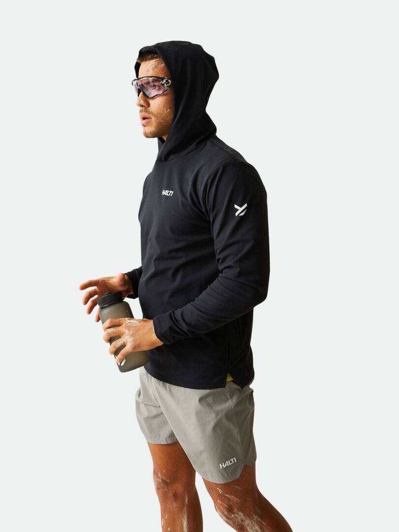 Eine Person in einem Halti Kaltio Trainingshoodie, grauen Shorts und Sonnenbrille hält eine Wasserflasche vor einem schlichten Hintergrund.