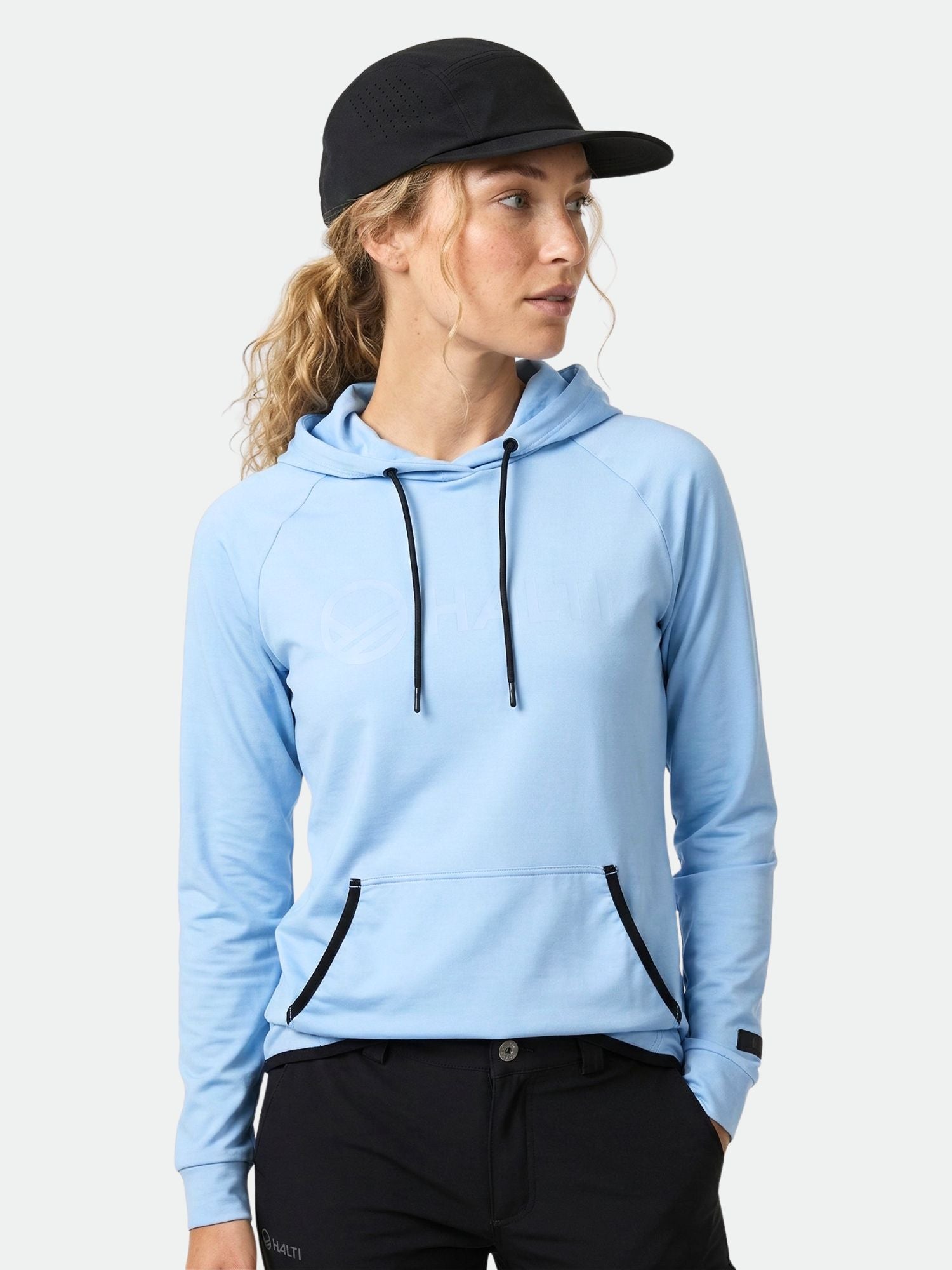 Eine Person trägt den Halti Veidnes Damen Hoodie in Hellblau, schwarze Mütze und Hose vor einem unifarbenen Hintergrund.