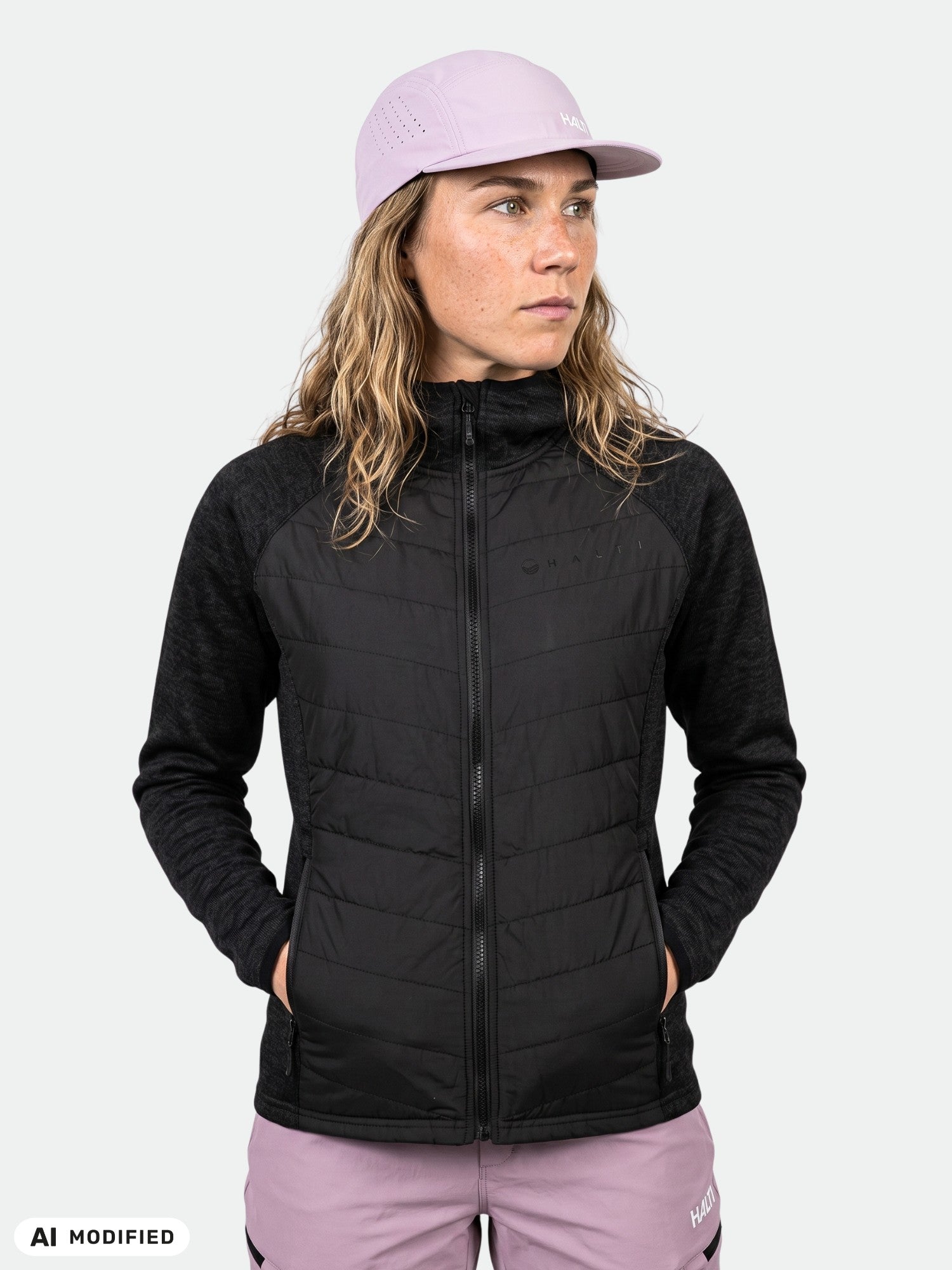 Frau in einer „Halti Streams Hybrid Midlayer“-Jacke und einer helllila Mütze, die Hände in den Taschen, vor einem neutralen Hintergrund.