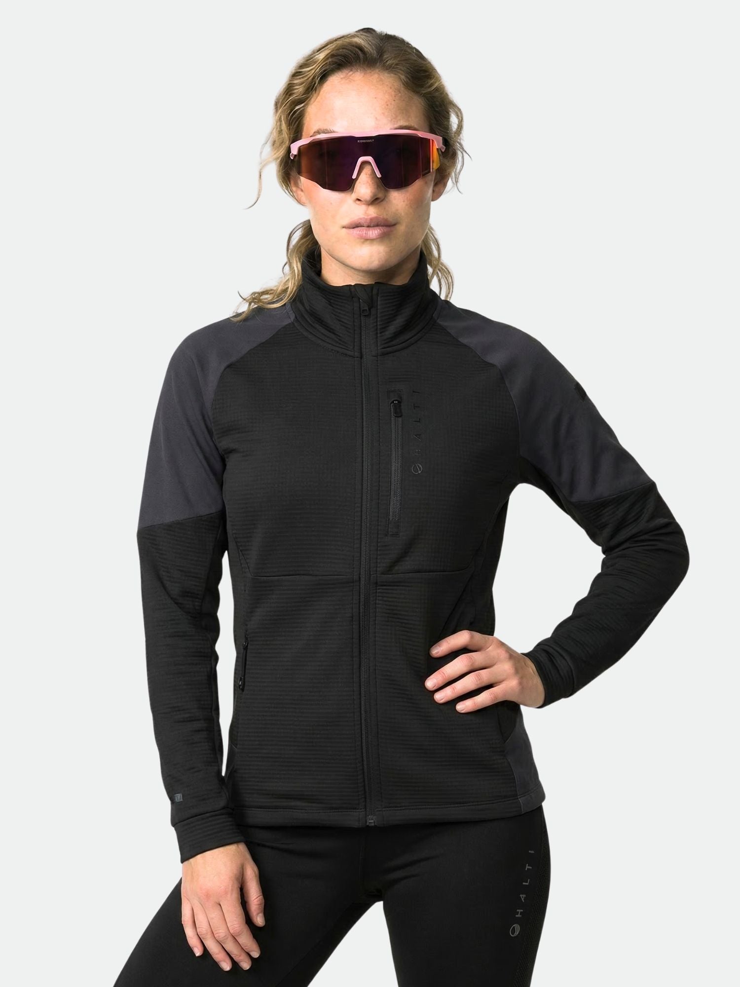 Eine Frau in einer schwarzen Halti Forerunner Damen Midlayer Jacke und Leggings posiert selbstbewusst mit sportlicher Sonnenbrille.