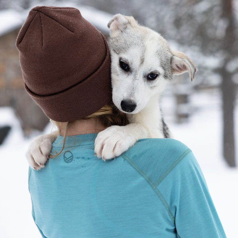 Eine Person mit einer braunen Mütze und einem Halti Pihka II Merinowolle Shirt Damen hält im Schnee einen Hund über der Schulter.