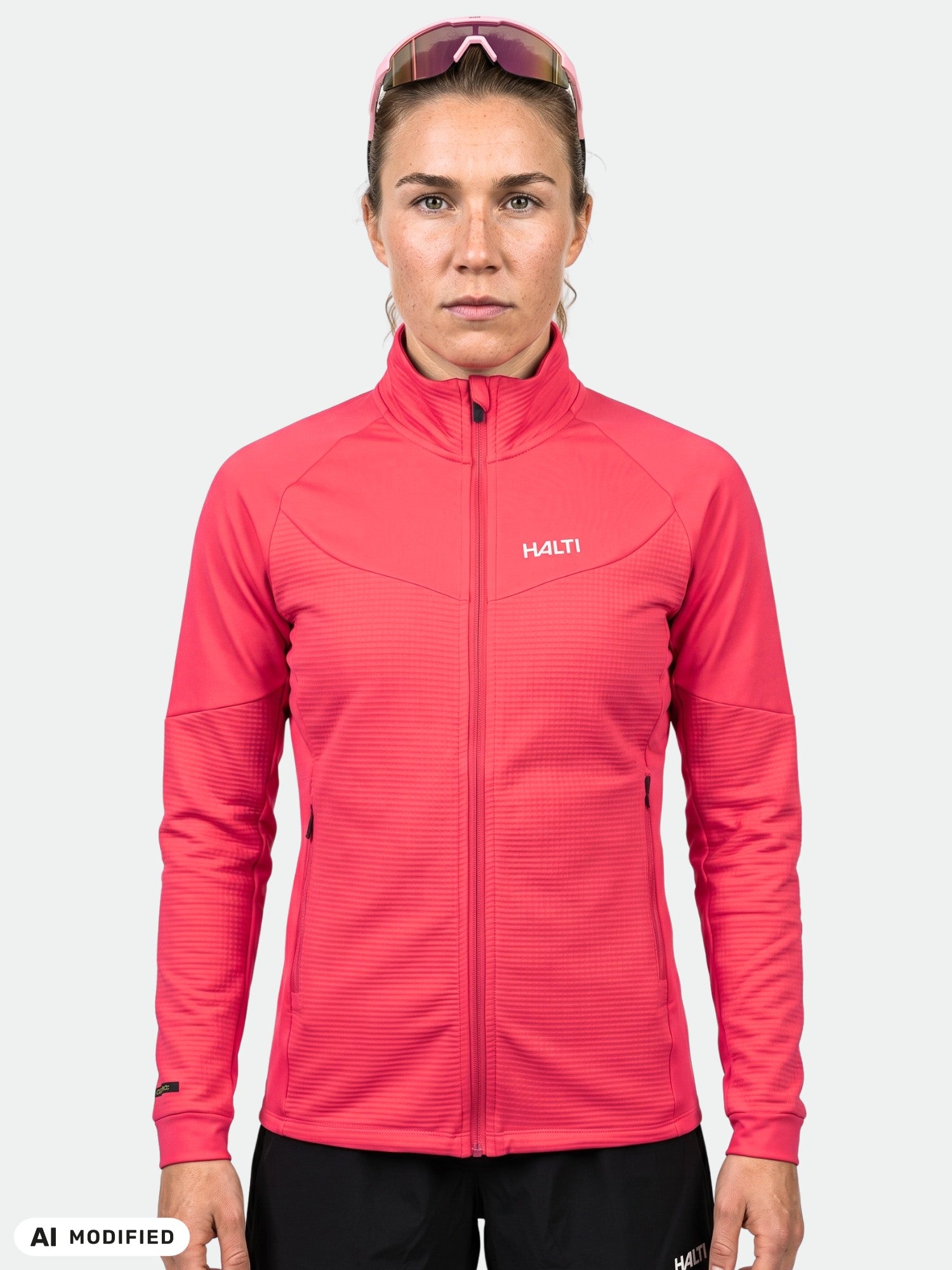 Eine Frau in einer leuchtend pinkfarbenen Halti HeatGrid Midlayer-Jacke und einer Sportsonnenbrille steht vor einem weißen Hintergrund.