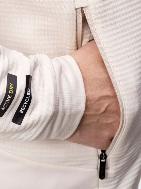 Eine Hand steckt in der Reißverschlusstasche einer cremefarbenen Halti HeatGrid Midlayer Jacke Damen mit Active Dry Tags.