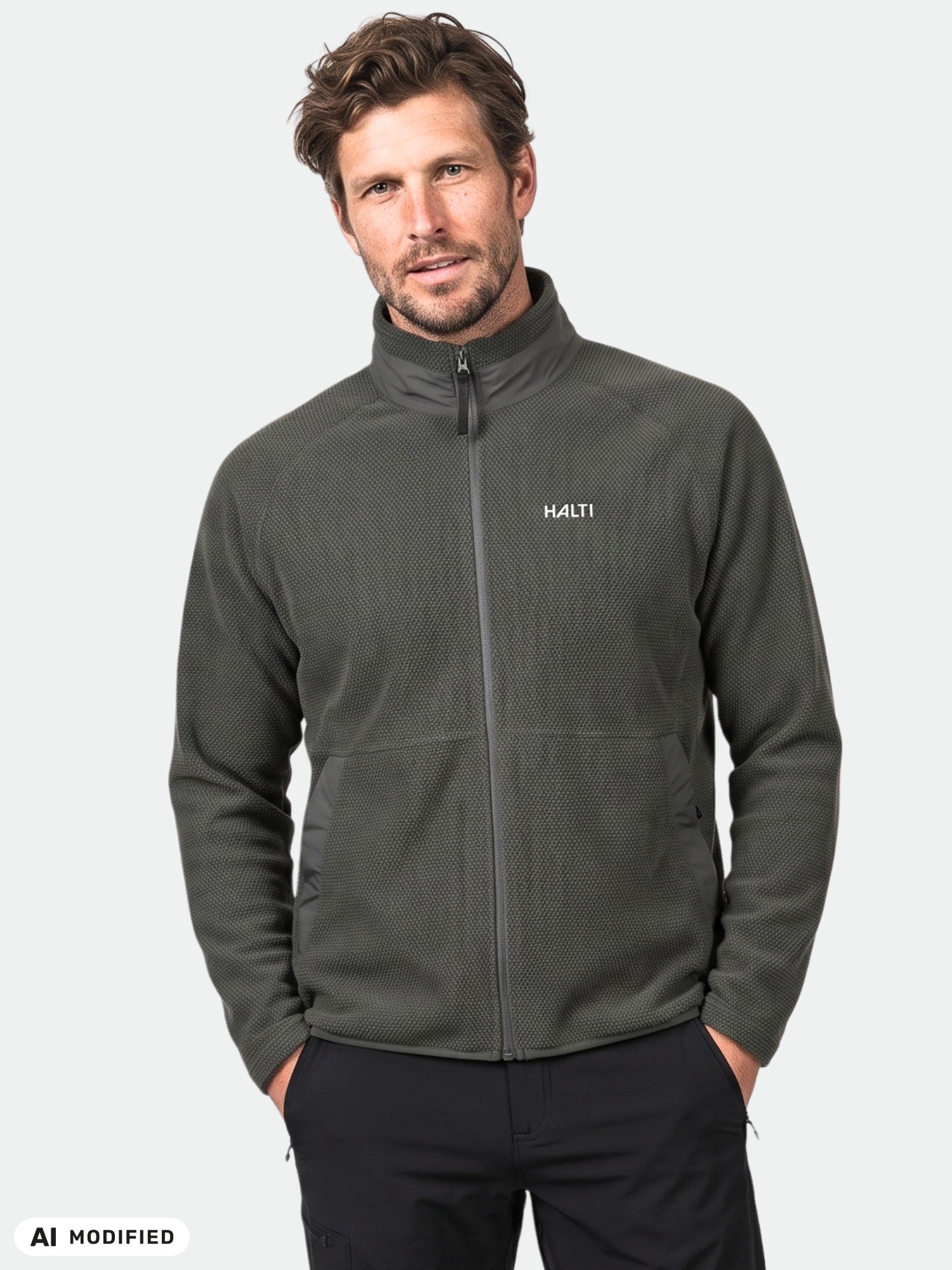 Ein Mann steht mit dem Blick nach vorne in einer dunklen Halti Soolo Midlayer Jacke für Herren, die Hände in den Taschen, vor einem einfarbigen Hintergrund.