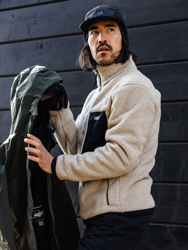 Mann in einer Halti Lumista Polar Fleece Jacke Herren und schwarzer Mütze hält einen grünen Mantel an eine dunkle Holzwand.