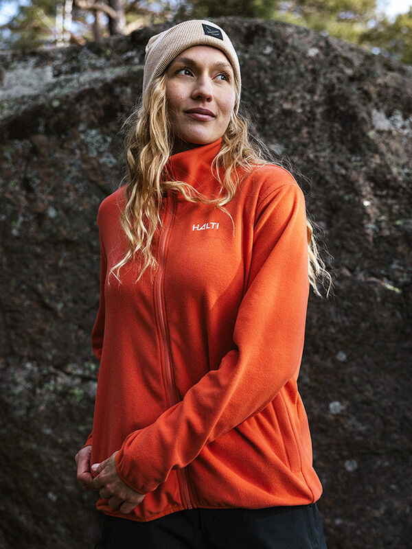 Eine Person in einer Halti Rockmoon Fleece Jacke Damen und beiger Mütze steht draußen vor einem großen Felsen.