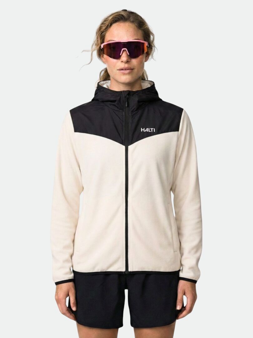 Person trägt Halti Rockmoon Fleecejacke Damen in creme/schwarz, dunkle Shorts und rosa Sonnenbrille vor einem unifarbenen Hintergrund.