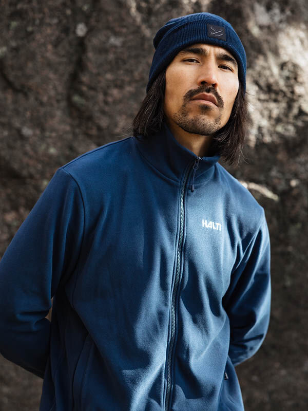 Ein Mann in einer Halti Rockmoon Fleecejacke Herren und Mütze steht an einem Felsen, bereit für Outdoor-Aktivitäten.