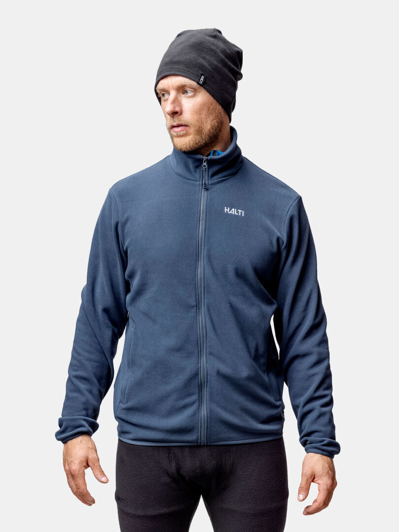 Ein Mann mit einer grauen Mütze, einer schwarzen Hose und einer Halti Rockmoon Fleece Jacke Herren steht vor einem hellen Hintergrund.