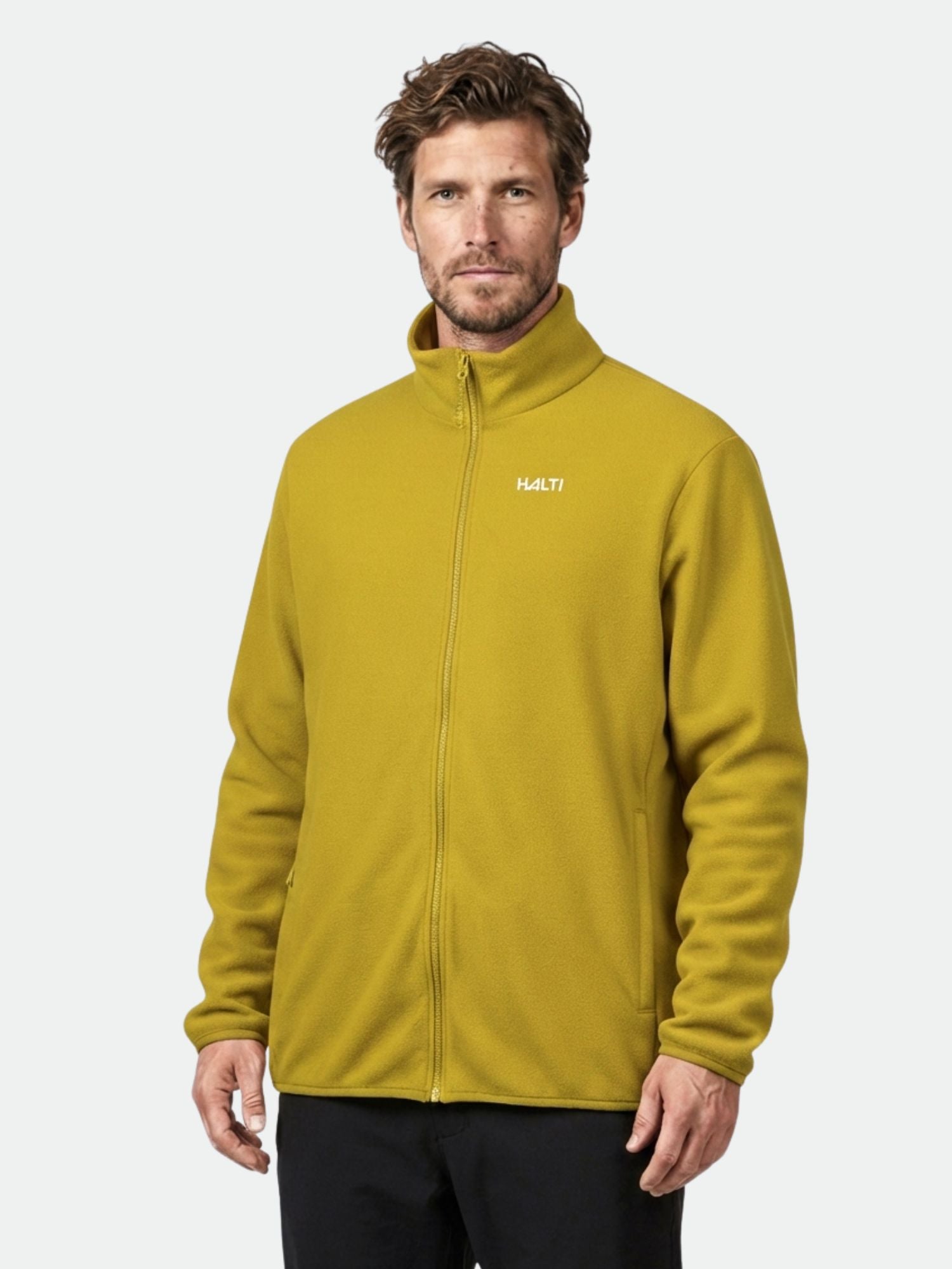 Ein Mann in einer senfgelben Halti Rockmoon Fleece Jacke Herren, perfekt als Midlayer für Outdoor-Aktivitäten, steht vor einem schlichten Hintergrund.