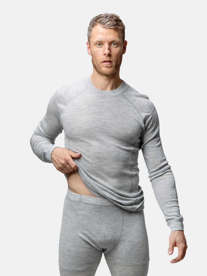 Ein Mann in Halti Hossa II Merino Unterwäsche Set Herren hebt sein Hemd, um seine Taille vor einem weißen Hintergrund zu zeigen.