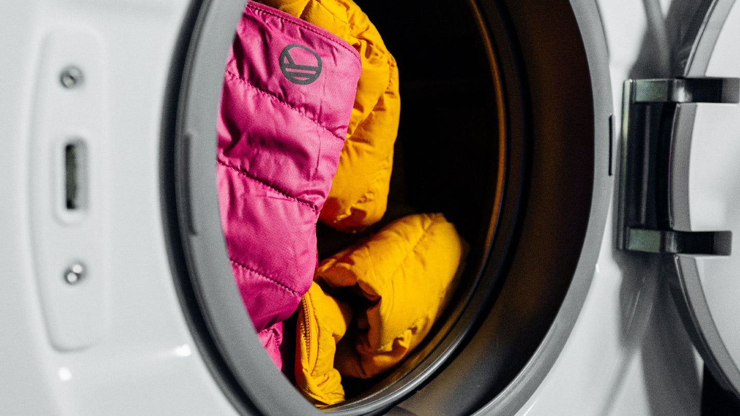 Eine Waschmaschine mit geöffneter Tür, in deren Trommel sich eine rosa und eine gelbe Jacke befinden.