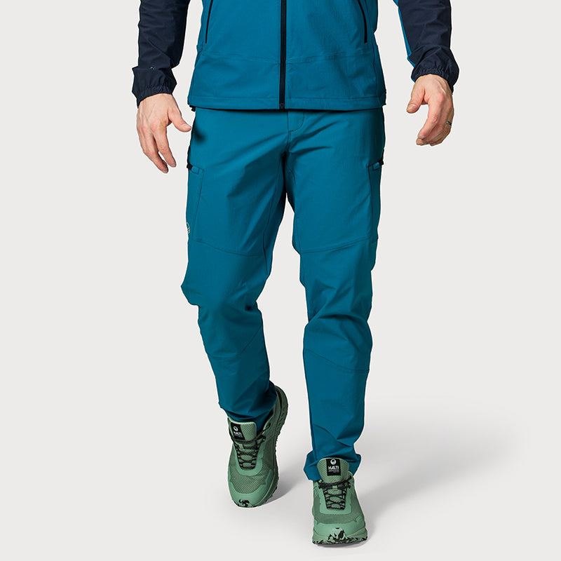 Eine Person in Halti Pallas Evo X-Stretch Hosen Herren, einer passenden Jacke und grünen Wanderschuhen, von den Knien bis zu den Füßen zu sehen.