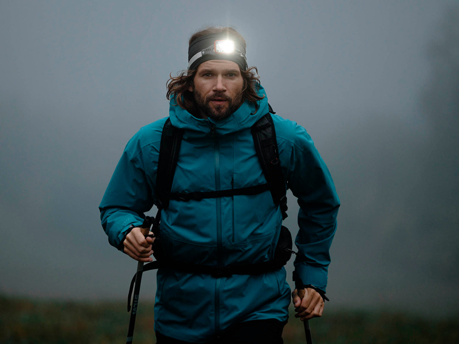 Eine Person in einer blauen Jacke wandert im Nebel, trägt eine Stirnlampe und hat Trekkingstöcke und einen Rucksack dabei.