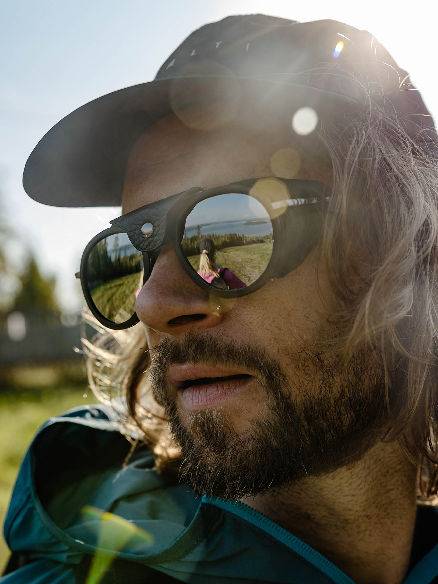 Ein Mann mit Bart und langen Haaren trägt eine Sonnenbrille und eine Mütze; in seiner Sonnenbrille spiegelt sich eine Landschaft.