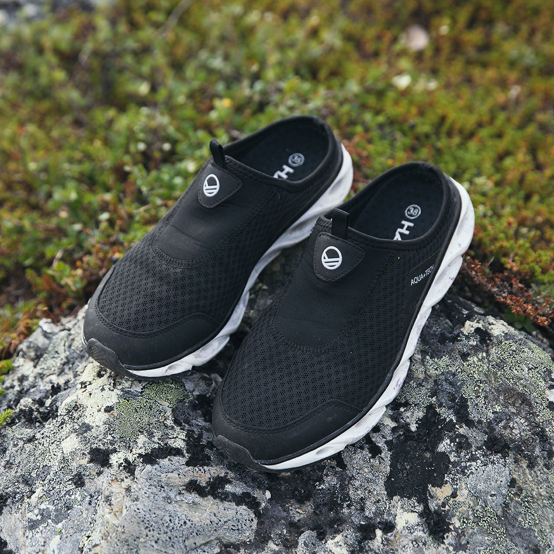 Ein Paar schwarze Mesh-Slipper mit weißen Sohlen, die auf einem Felsen mit grüner Vegetation im Hintergrund stehen.