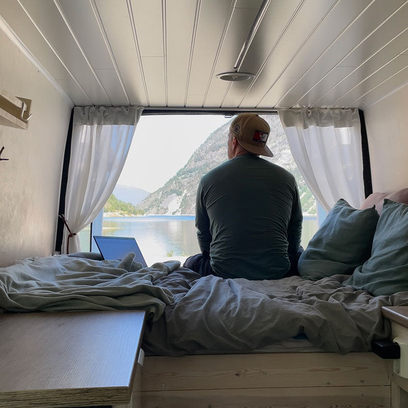 Eine Person sitzt auf einem Bett in einem Lieferwagen und blickt durch die offenen Hintertüren auf einen See und die Berge.