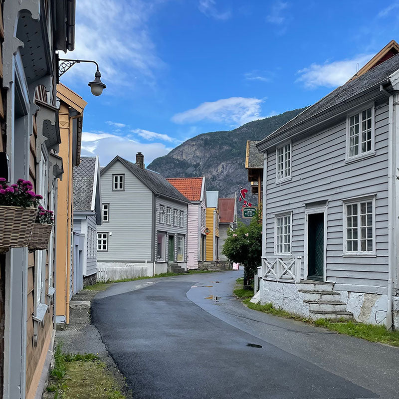 Enge, mit Holzhäusern gesäumte Straße, Berge im Hintergrund und ein teilweise bewölkter blauer Himmel darüber.