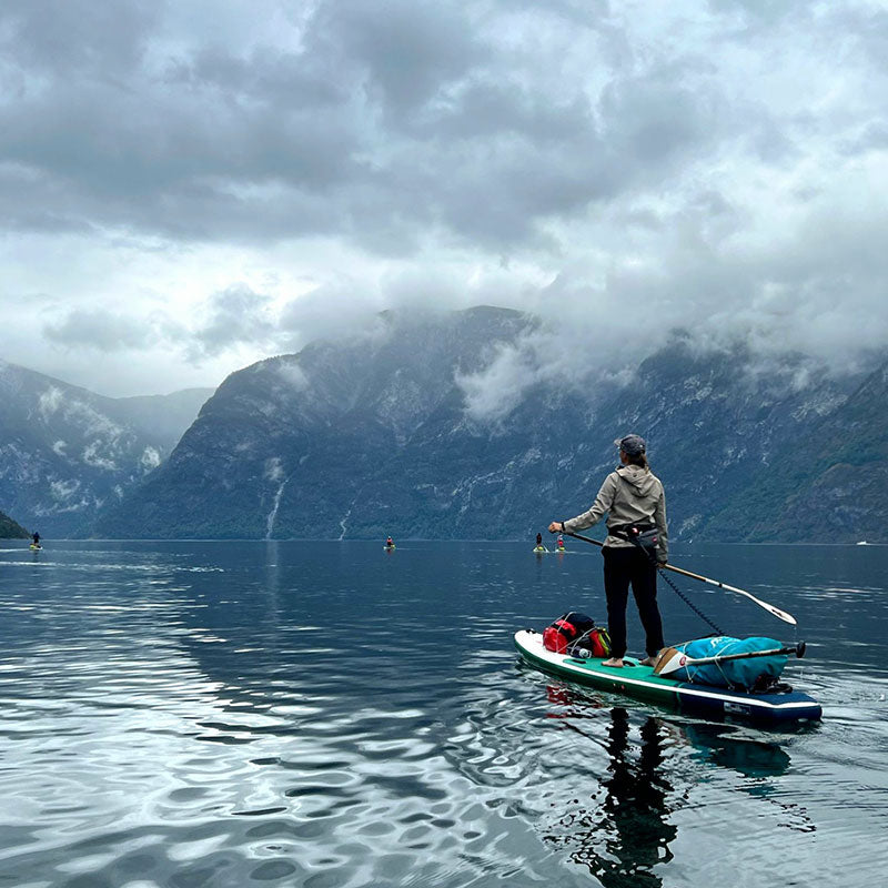 Eine Person steht auf einem Paddelbrett auf einem ruhigen See, umgeben von Bergen und Wolken unter einem bedeckten Himmel.