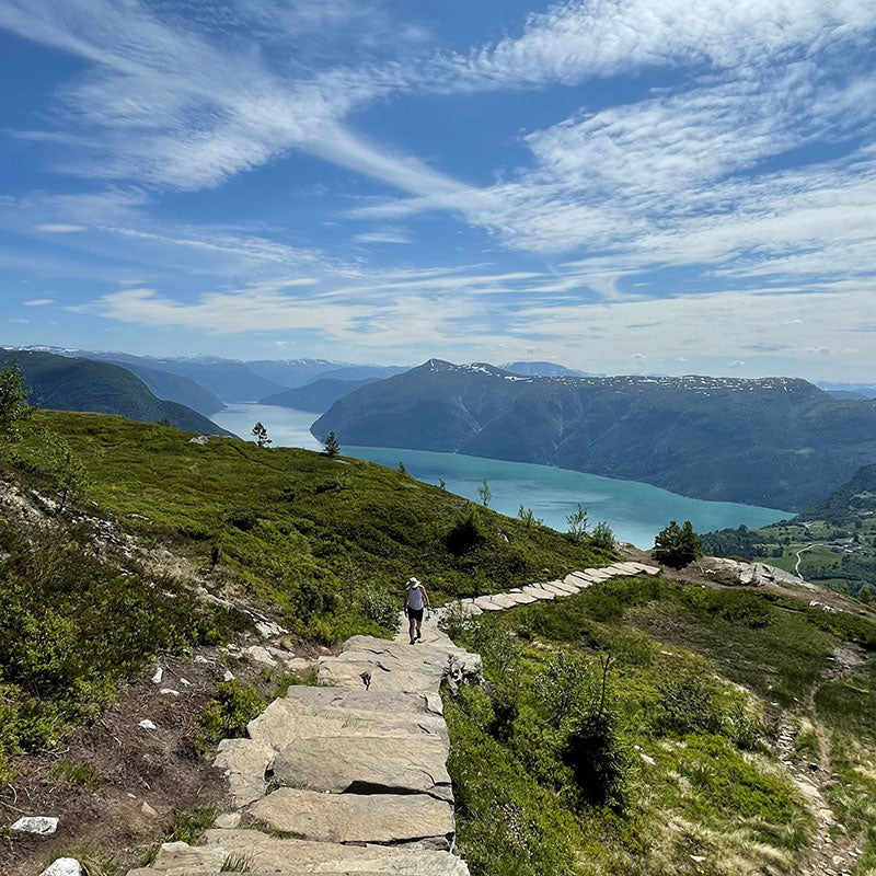 Eine Person geht einen steinernen Weg auf einem Berg hinunter, mit Blick auf einen See und ferne Hügel unter einem teilweise bewölkten Himmel.