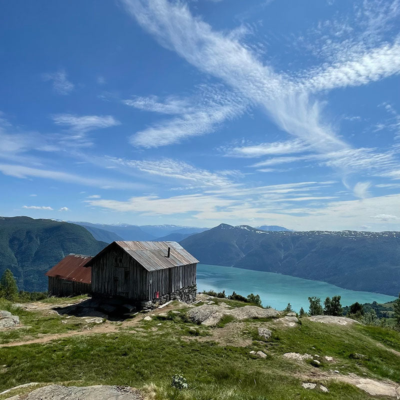 Eine verwitterte Holzhütte steht auf einer felsigen Anhöhe mit Blick auf einen See und ferne Berge unter einem blauen, wolkenverhangenen Himmel.