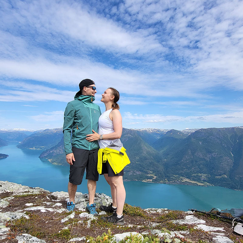 Zwei Personen stehen auf einem Felsvorsprung mit Blick auf einen blauen See und Berge unter einem teilweise bewölkten Himmel.