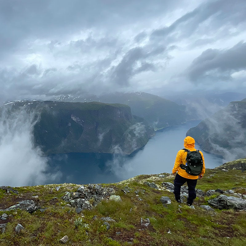 Person in gelber Jacke steht auf einer grasbewachsenen Klippe mit Blick auf einen nebligen Fjord und Berge unter einem bewölkten Himmel.