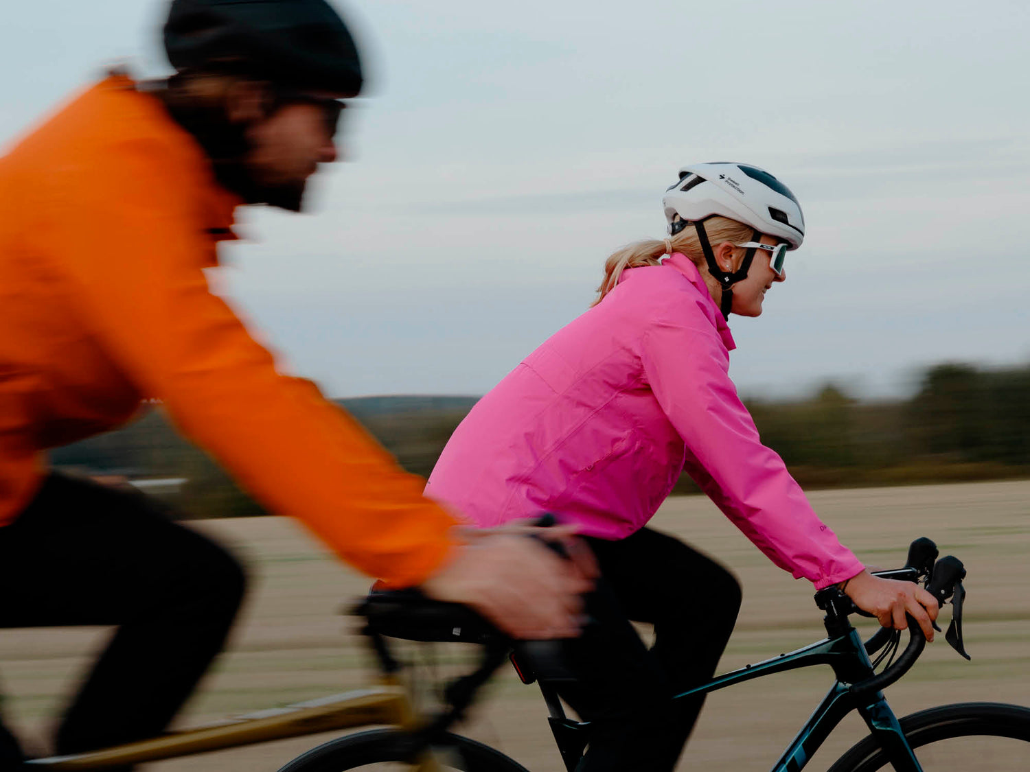 Zwei Radfahrer mit Helmen und Jacken fahren im Freien nebeneinander, einer in Orange, der andere in Rosa.