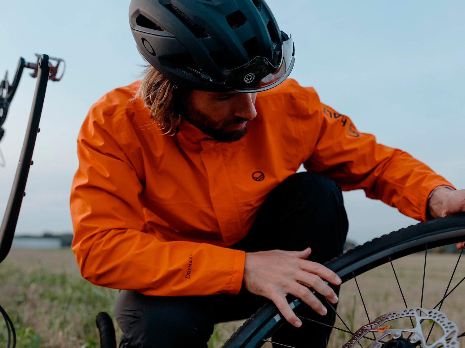 Eine Person in einer orangefarbenen Jacke und mit Helm inspiziert einen Fahrradreifen auf einer Wiese.