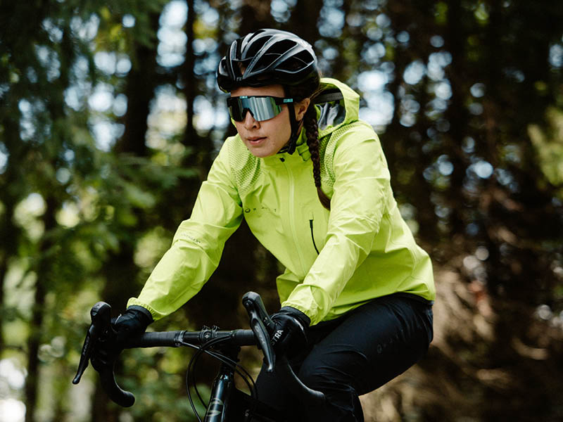 Eine Person mit neongelber Jacke, Helm und Sonnenbrille fährt mit dem Fahrrad im Freien durch ein bewaldetes Gebiet.