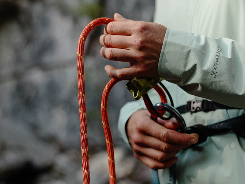 Eine Person, die eine leichte Jacke trägt, hält ein Kletterseil und ein Sicherungsgerät und bereitet sich auf das Klettern im Freien vor.