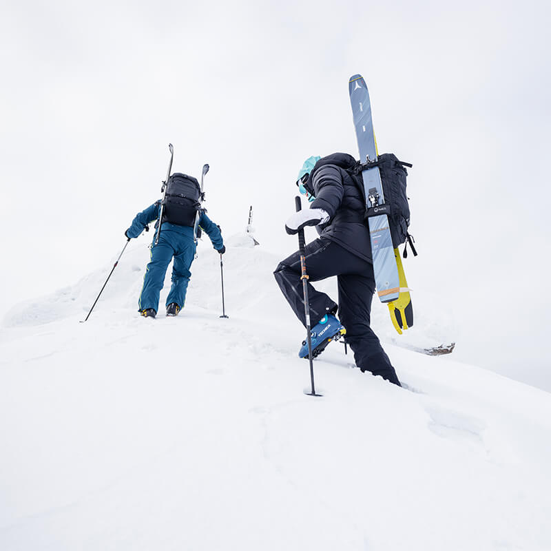 Zwei Personen in Winterkleidung wandern bei bedecktem Himmel mit Skiern im Rucksack bergauf.