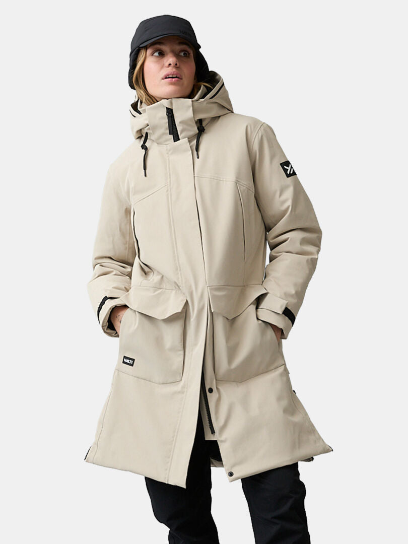 Frau in einem beigen Halti Kallio DX Parka, schwarzer Hose und Mütze, die Hände in den Taschen, vor einfarbigem Hintergrund.