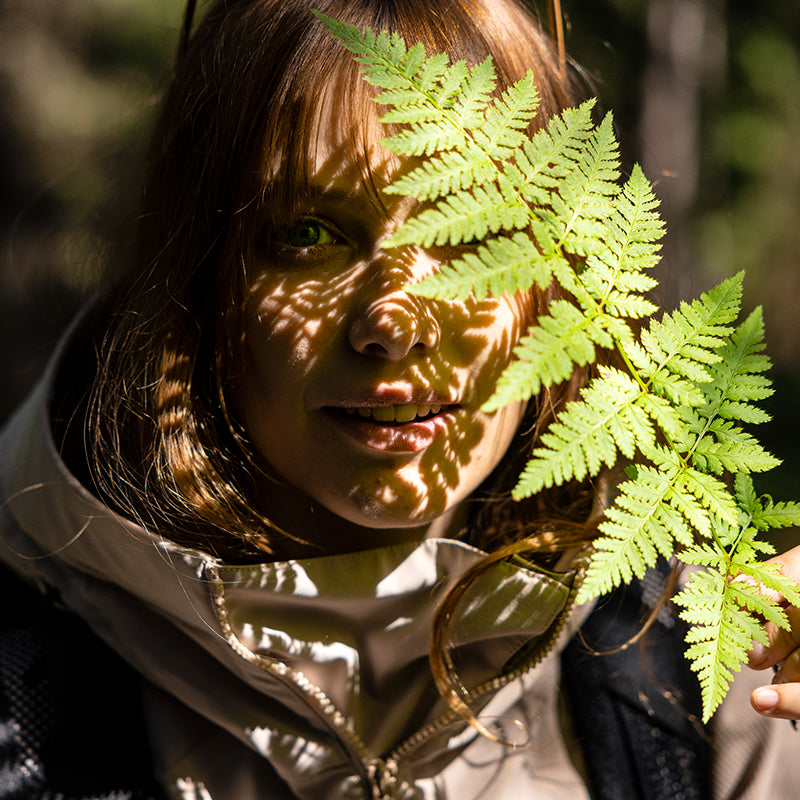 Eine Person hält ein grünes Farnblatt, das im Sonnenlicht ein Schattenmuster auf ihr Gesicht wirft.